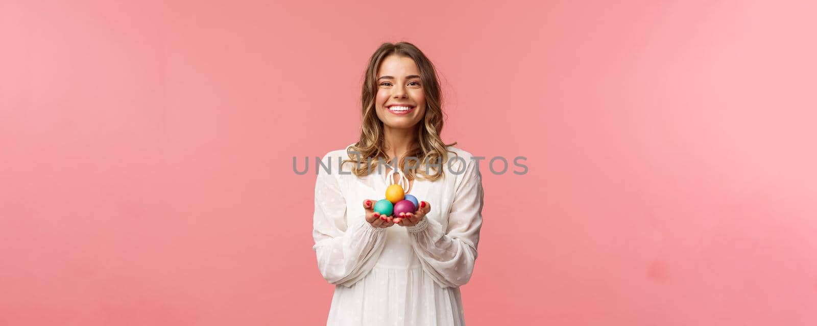 Holidays, spring and party concept. Portrait of tender, lovely blonde girl in white dress, holding Easter painted eggs, celebrating orthodox day, smiling cheerful share positivity, pink background.