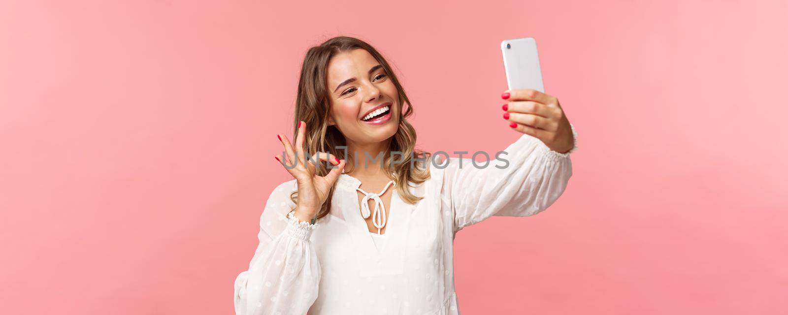 Close-up of satisfied good-looking blond girl in white dress, taking selfie, record mobile phone video, show okay satisfactory sign with pleased nod, smiling agree or recommend, pink background.