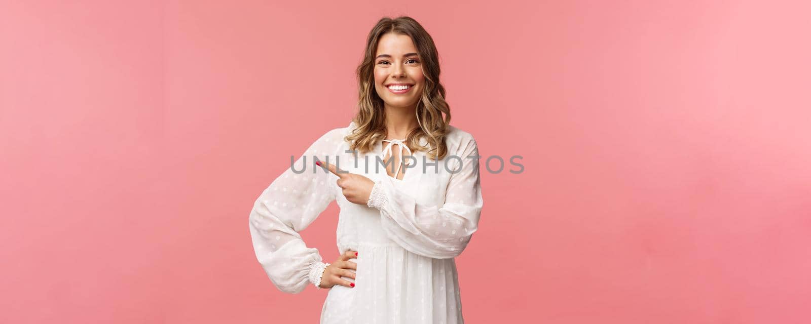 Pleasant good-looking smiling blond woman with blond short wavy hair, white tender dress, looking camera and pointing finger left to show you best price, good offer, promo concept, pink background.