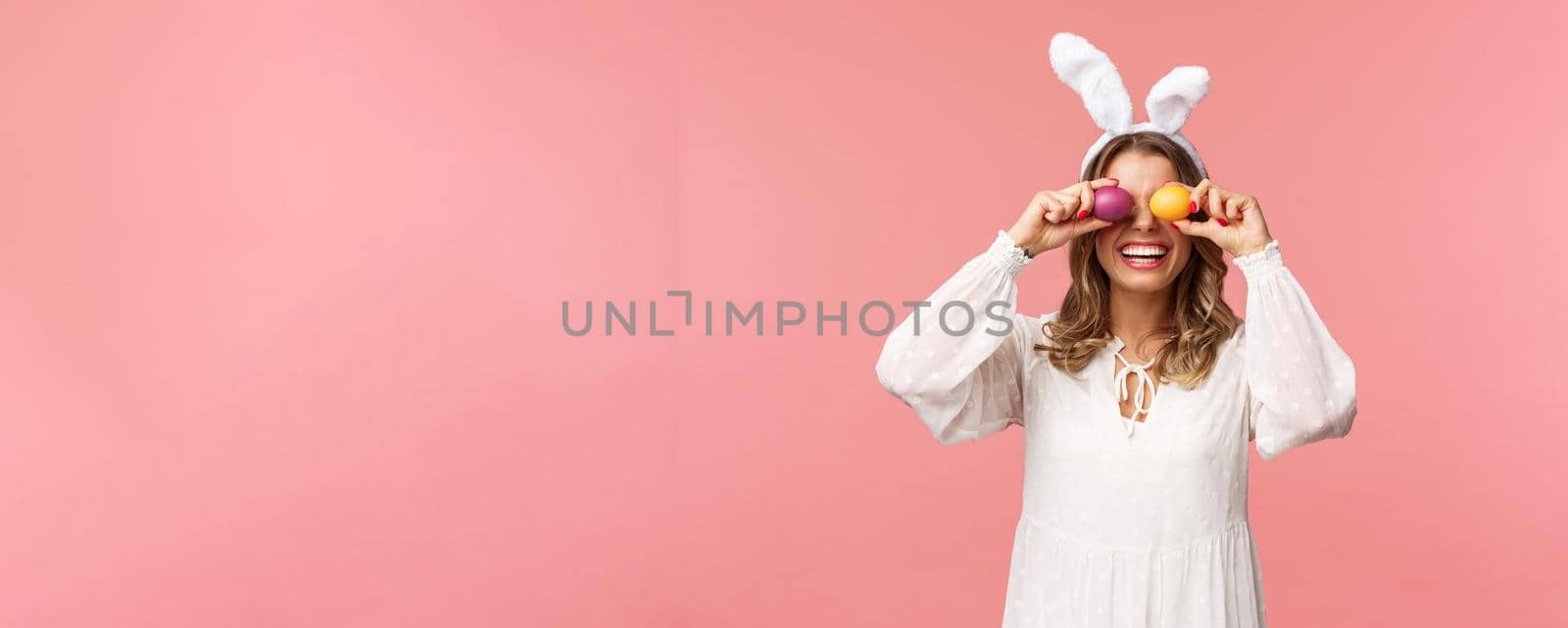 Holidays, spring and party concept. Portrait of lovely, tender smiling woman in rabbit ears and white dress celebrating Easter day, holding painted eggs on eyes and grinning, pink background.