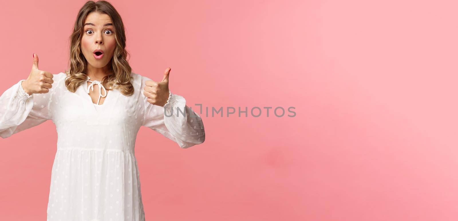 Portrait of amazed, excited blond young girl in white cute dress, open mouth say wow astonished and fascinated, show thumbs-up like and approve, beaing satisfied, pink background by Benzoix