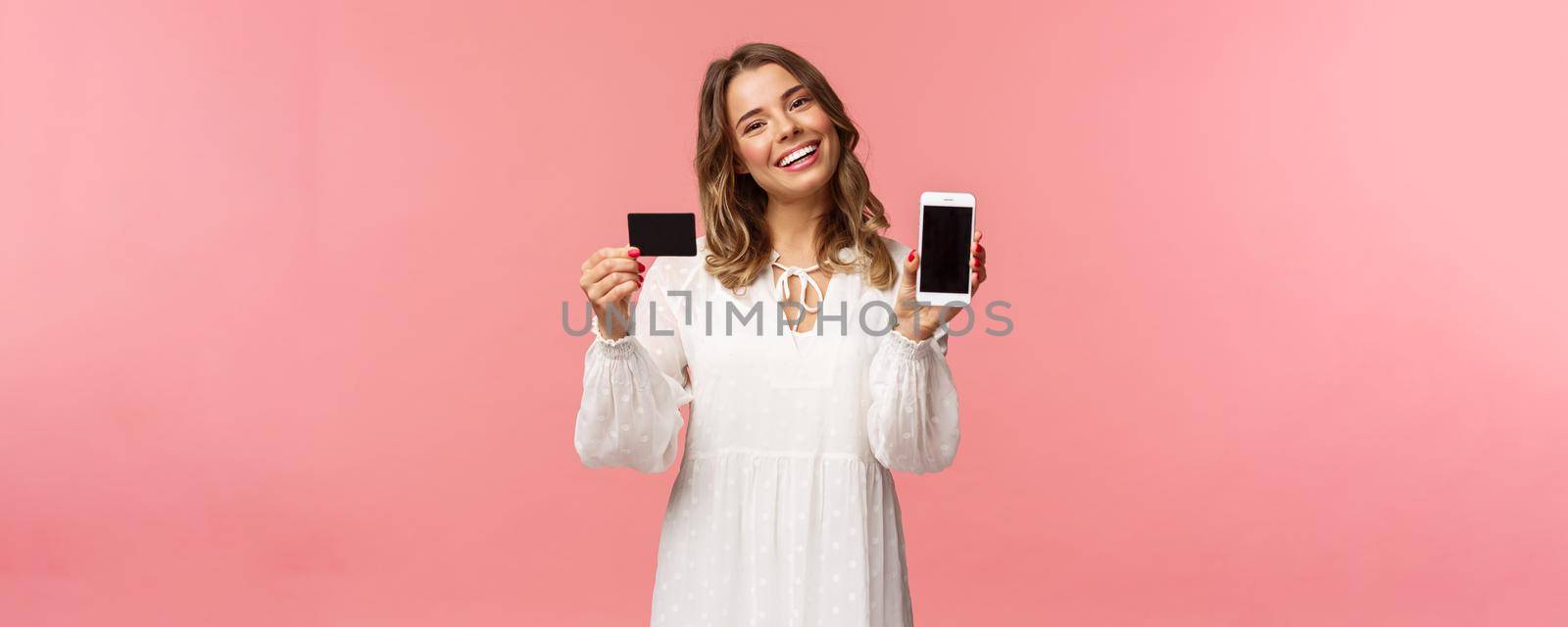 Portrait of tender feminine young blond girl in white dress, tilt head and smiling pleased, advice download application, holding mobile phone, credit card, show smartphone screen, pink background.