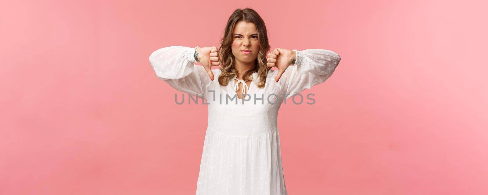 Portrait of picky young blond caucasian woman in white dress over pink background expressing dislike, show thumbs-down and grimacing in aversion, disappointed with bad quality.