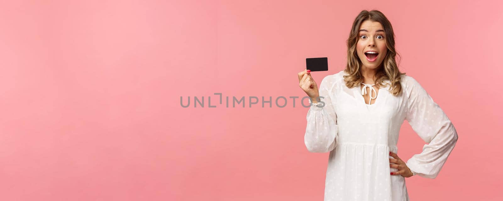 Portrait of happy excited good-looking blond girl in white dress, being impressed and pleased with cool banking service, showing credit card, smiling camera, standing pink background by Benzoix