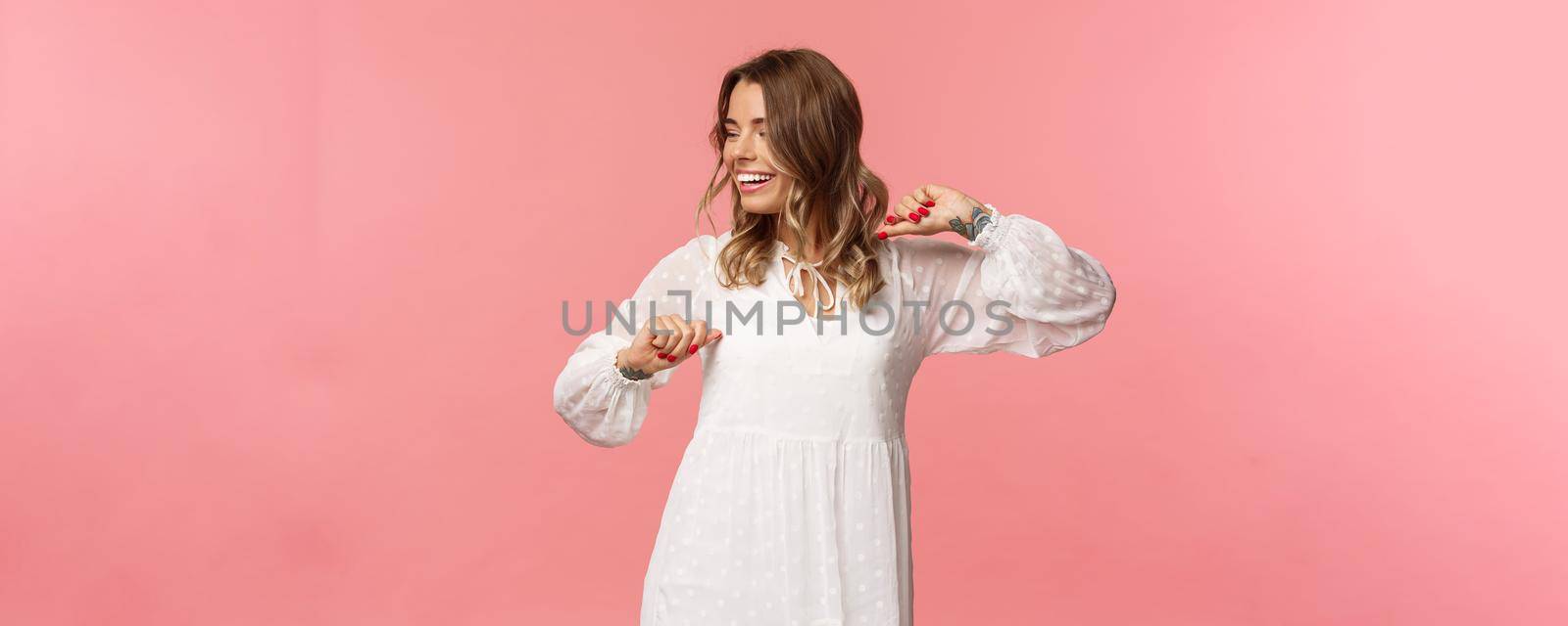 Beauty, fashion and women concept. Tender and carefree pretty young girl enjoying spring time, wearing white dress, dancing and looking away with beaming smile having fun, pink background.