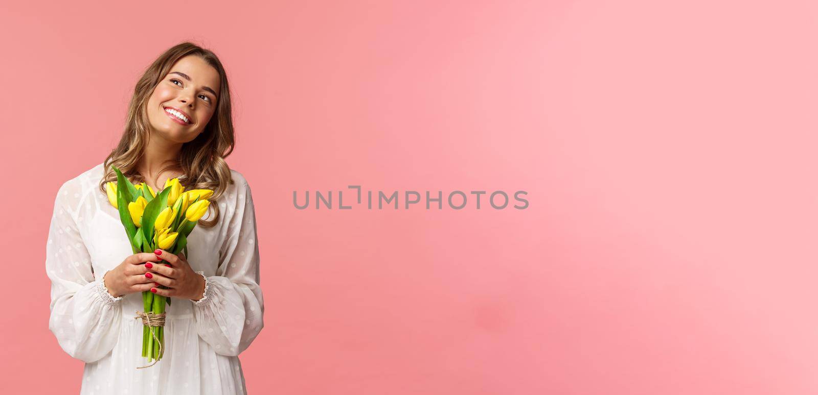 Holidays, beauty and spring concept. Portrait of dreamy, happy blond girl feeling romantic daydreaming about her girlfriend, holding yellow tulips, wear white dress, standing pink background by Benzoix