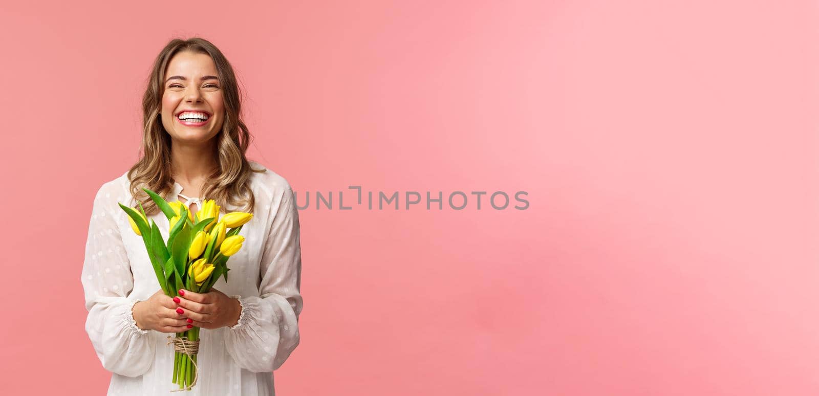 Holidays, beauty and spring concept. Portrait of happy excited charming blond girl receive flowers, buying yellow tulips herself, smiling and laughing joyfully, stand pink background.