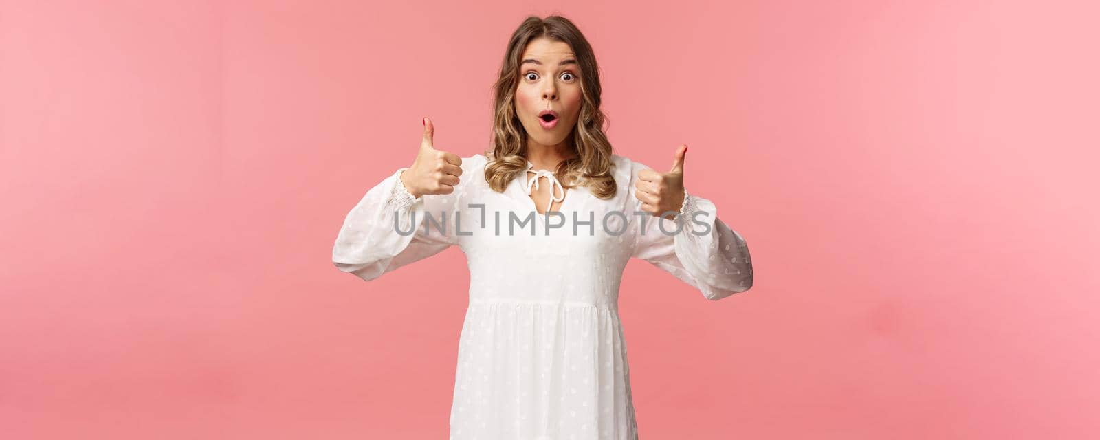 Portrait of amazed, excited blond young girl in white cute dress, open mouth say wow astonished and fascinated, show thumbs-up like and approve, beaing satisfied, pink background.