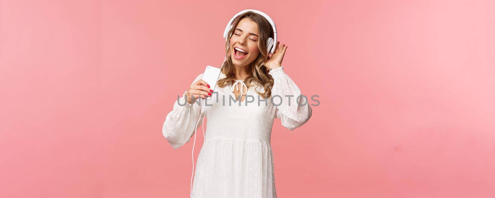 Portrait of beautiful blond caucasian female in white dress, listening music in headphones, close eyes and smiling, using mobile phone like microphone, singing along favorite song, pink background by Benzoix