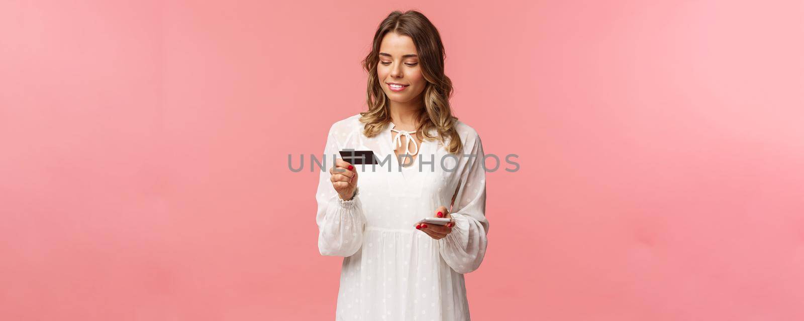 Portrait of attractive and cute blond girl with short curly hairstyle, white dress, holding smartphone and credit card, insert digit numbers to register in shopping app, buy online, pink background.