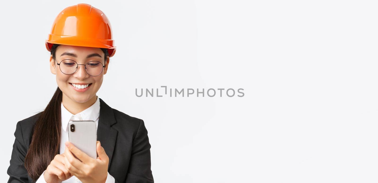 Close-up of smiling professional asian female industrial engineer, wearing safety helmet and business suit, using smartphone, looking pleased at mobile phone screen, standing white background.