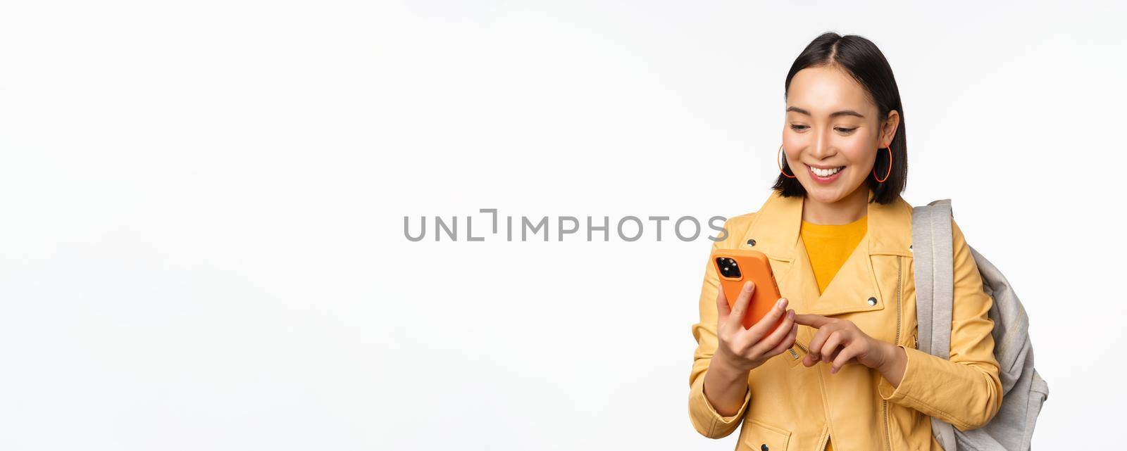 Image of happy girl traveller, tourist with backpack looking at smartphone, using mobile phone route app, standing over white background. Copy space