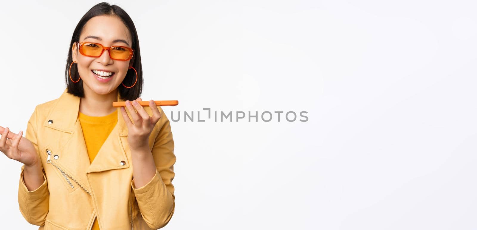 Image of happy asian girl talking on speakerphone, recording, translating her voice with mobile phone app, talking in smartphone dynamic, standing over white background.