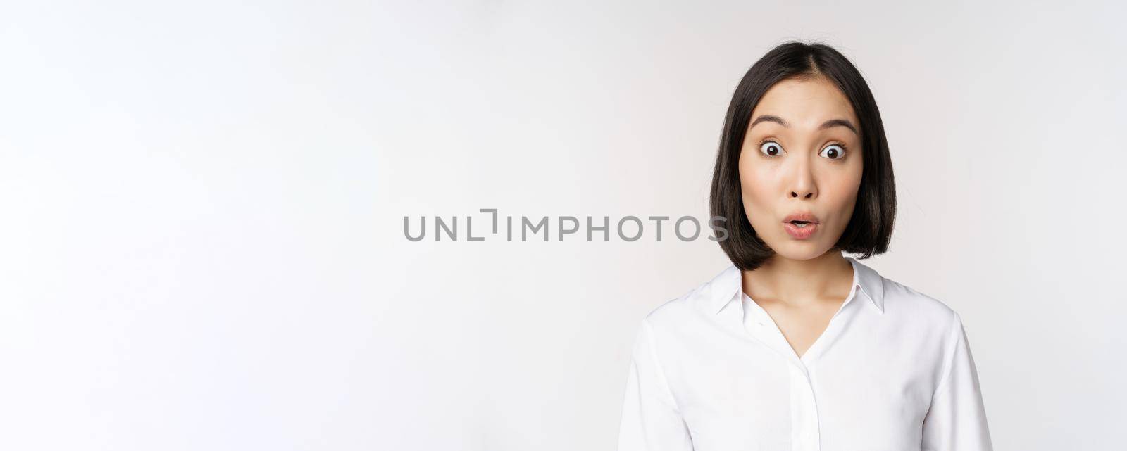 Close up portrait of young asian female model looking amazed at camera, smiling white teeth, standing against white background by Benzoix