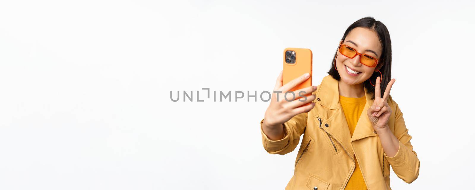 Stylish, beautiful asian girl in sunglasses, taking selfie on smartphone, posing for photo, holding mobile phone, white studio background.