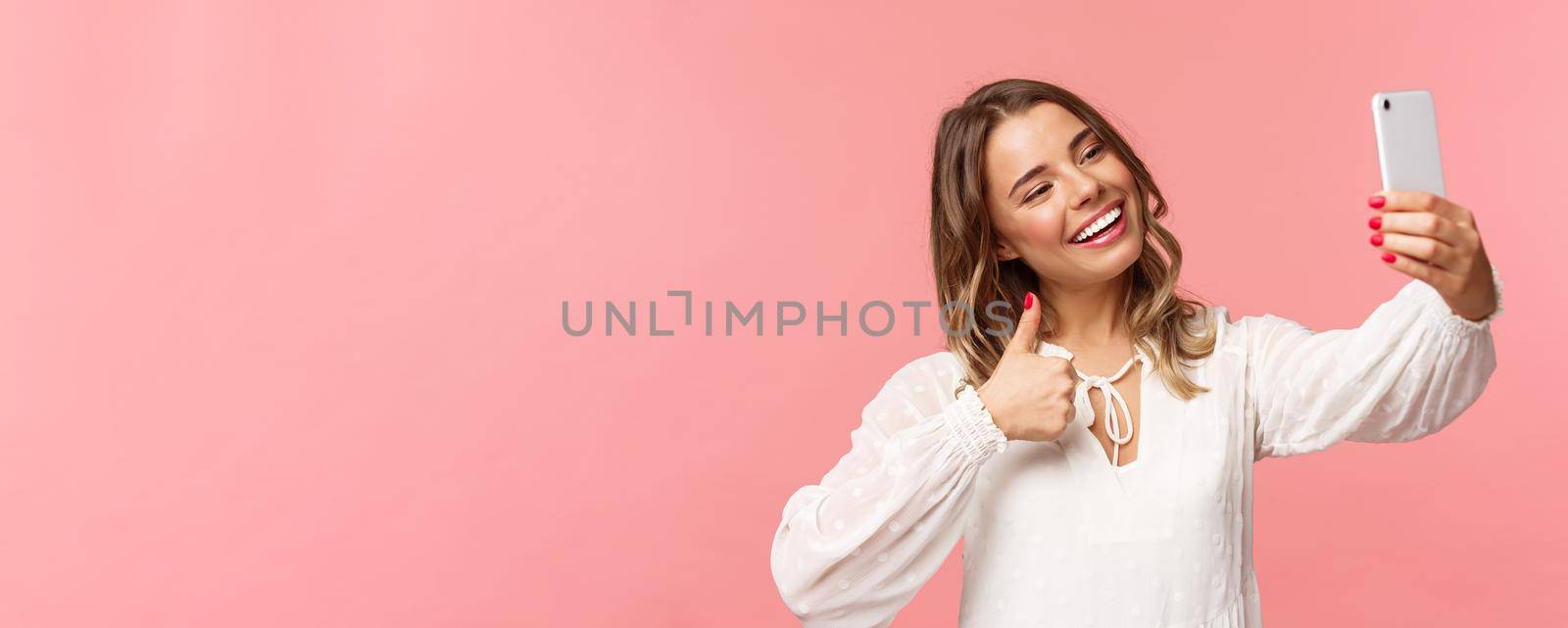 Close-up portrait of stylish beautiful beauty blogger record video on smartphone, taking selfie, showing thumbs-up at mobile phone camera, smiling pleased, recommend place to followers by Benzoix