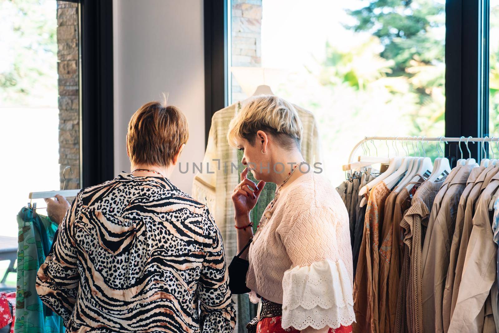 old woman with red hair, dressed in casual and modern way, mature woman blonde, dressed in long red skirt and pink jumper. two women in an exclusive fashion shop, buying clothes, mother and daughter enjoying a shopping day together. shopping concept. clothes seller in her small shop. natural light from window, sun rays, nature background plants, clothes racks, shop windows, clothes, Horizontal.