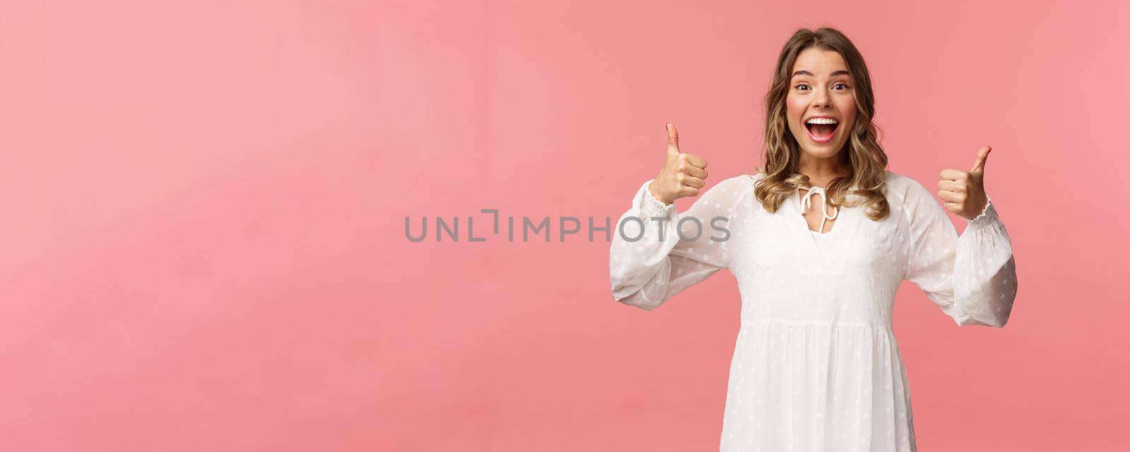 Portrait of excited feminine blond girl in white dress, show thumbs-up in approval, like being pleased, nod agreement, smiling fascinated, leave positive review, recommend product, pink background.