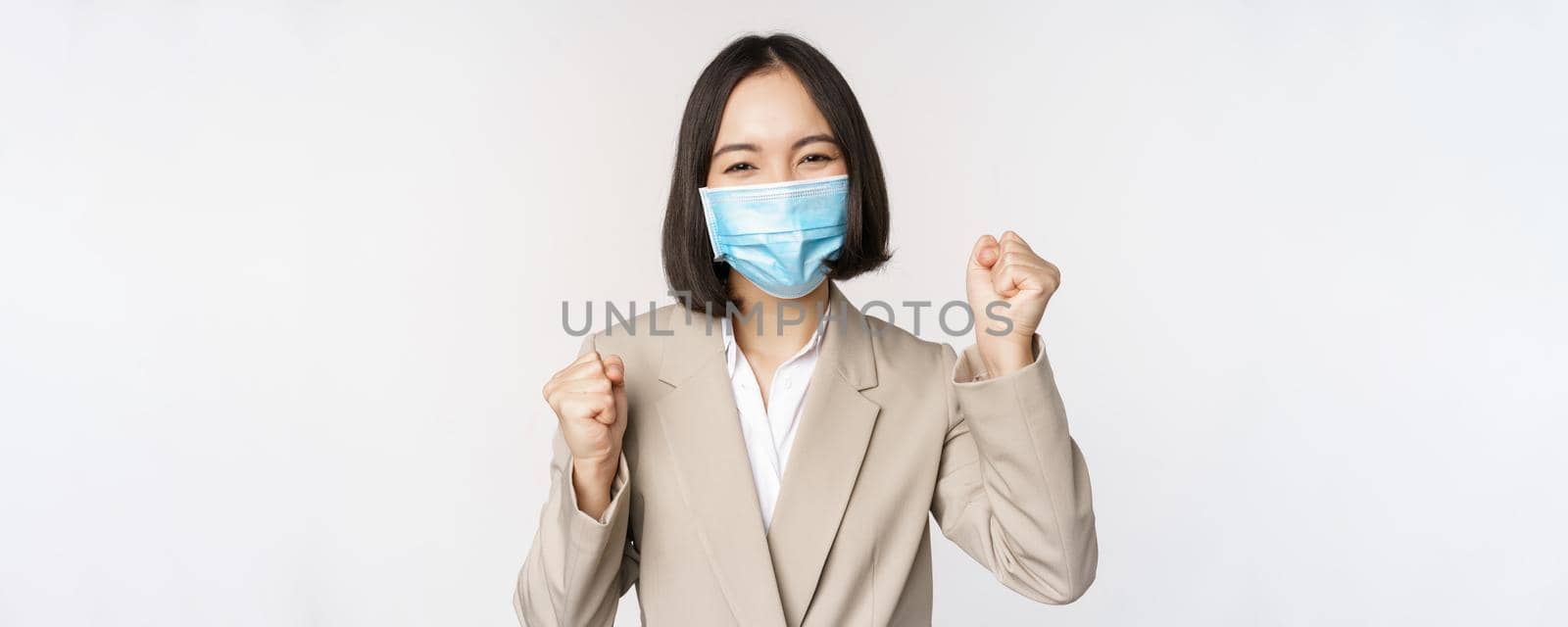 Coronavirus and business people concept. Happy businesswoman in medical face mask dancing, celebrating success, achievement, standing over white background by Benzoix