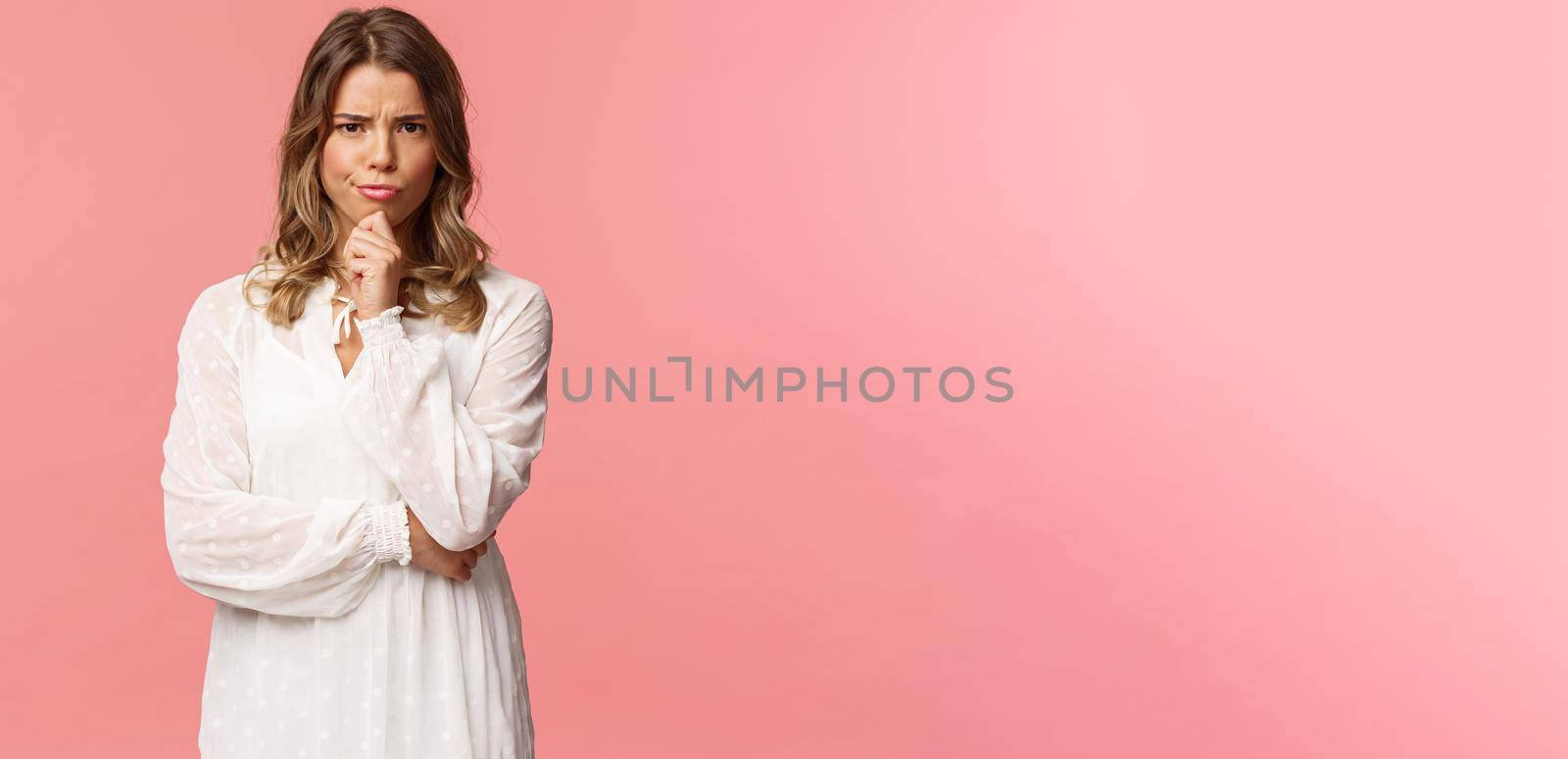 Beauty, fashion and women concept. Portrait of skeptical and judgemental serious-looking blond female in white spring dress, look disbelief, grimacing and frowning, pink background by Benzoix