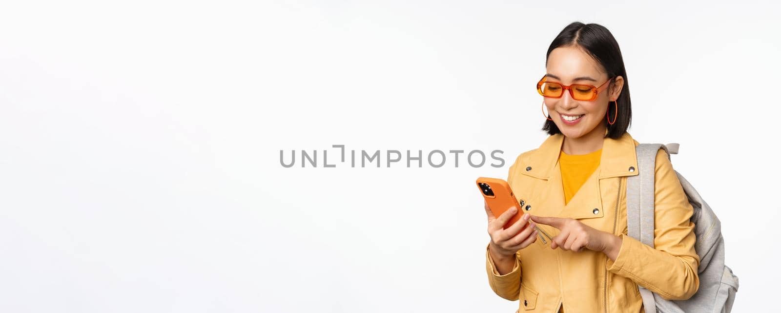 Stylish young asian woman tourist, traveller with backpack and smartphone smiling at camera, posing against white background by Benzoix