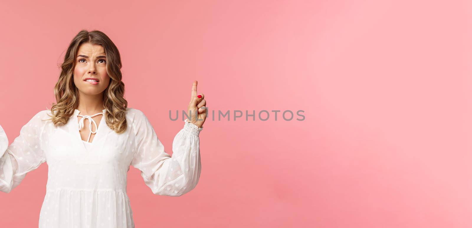 Skeptical and embarrassed blond caucasian woman in white cute dress, grimacing, cringe from seeing something disgusting, frowning doubtful and pointing fingers up strange thing, pink background.