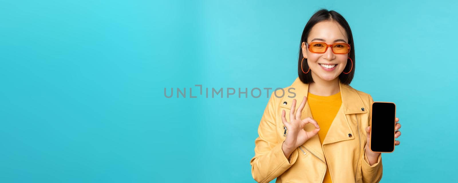 Smiling korean woman showing mobile phone app interface, smartphone application, recommending on cellphone, standing over blue background by Benzoix