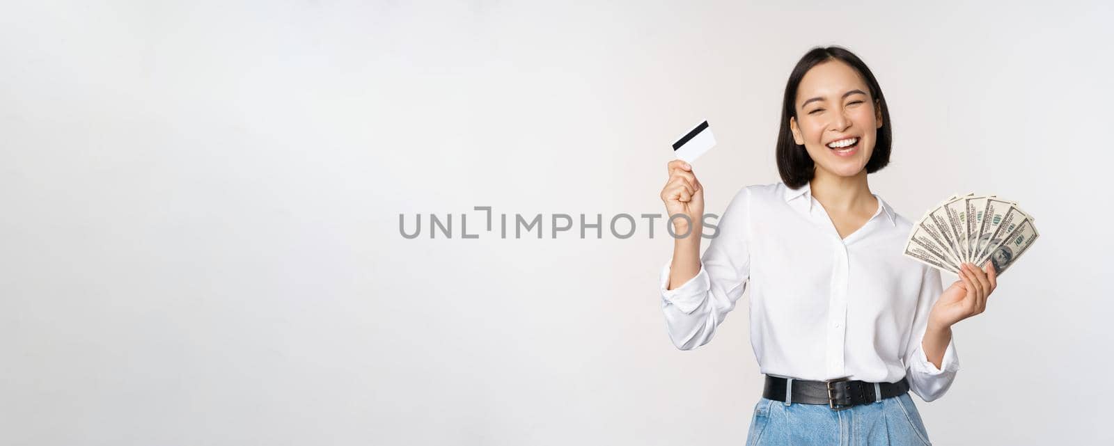 Happy korean woman holding credit card and money dollars, smiling and laughing, posing against white studio background by Benzoix