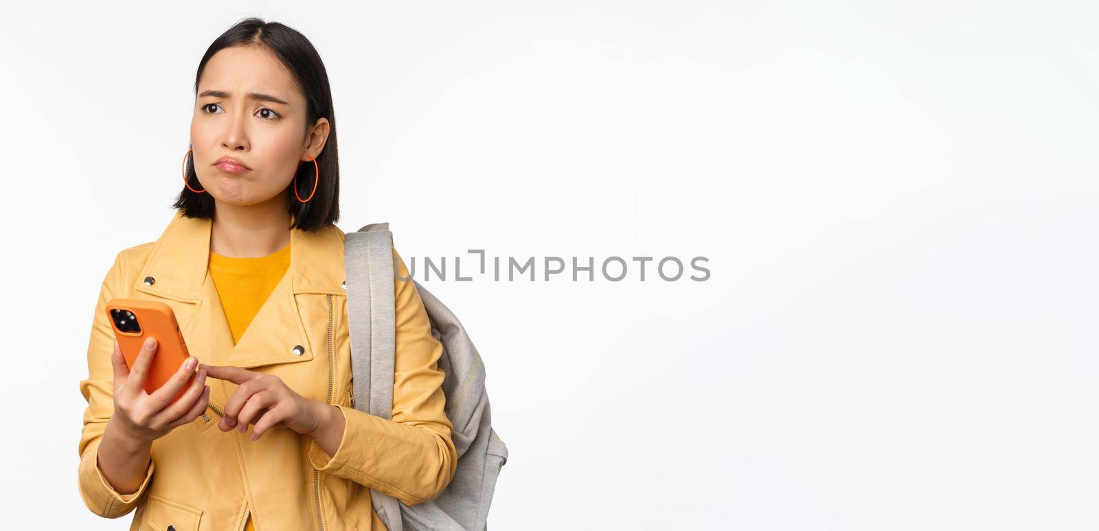 Image of happy girl traveller, tourist with backpack looking at smartphone, using mobile phone route app, standing over white background. Copy space