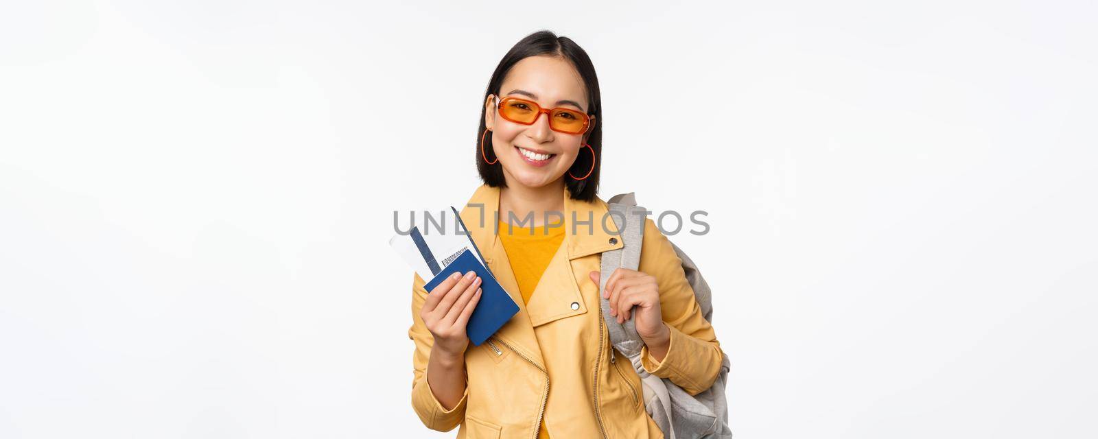 Happy asian girl going on vacation, holding passport and flight tickets, backpack on shoulder. Young woman tourist travelling abroad, standing over white background by Benzoix