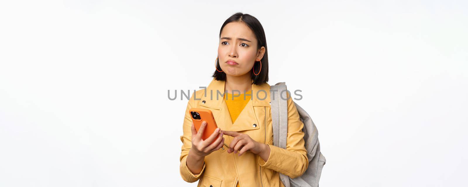 Image of happy girl traveller, tourist with backpack looking at smartphone, using mobile phone route app, standing over white background by Benzoix