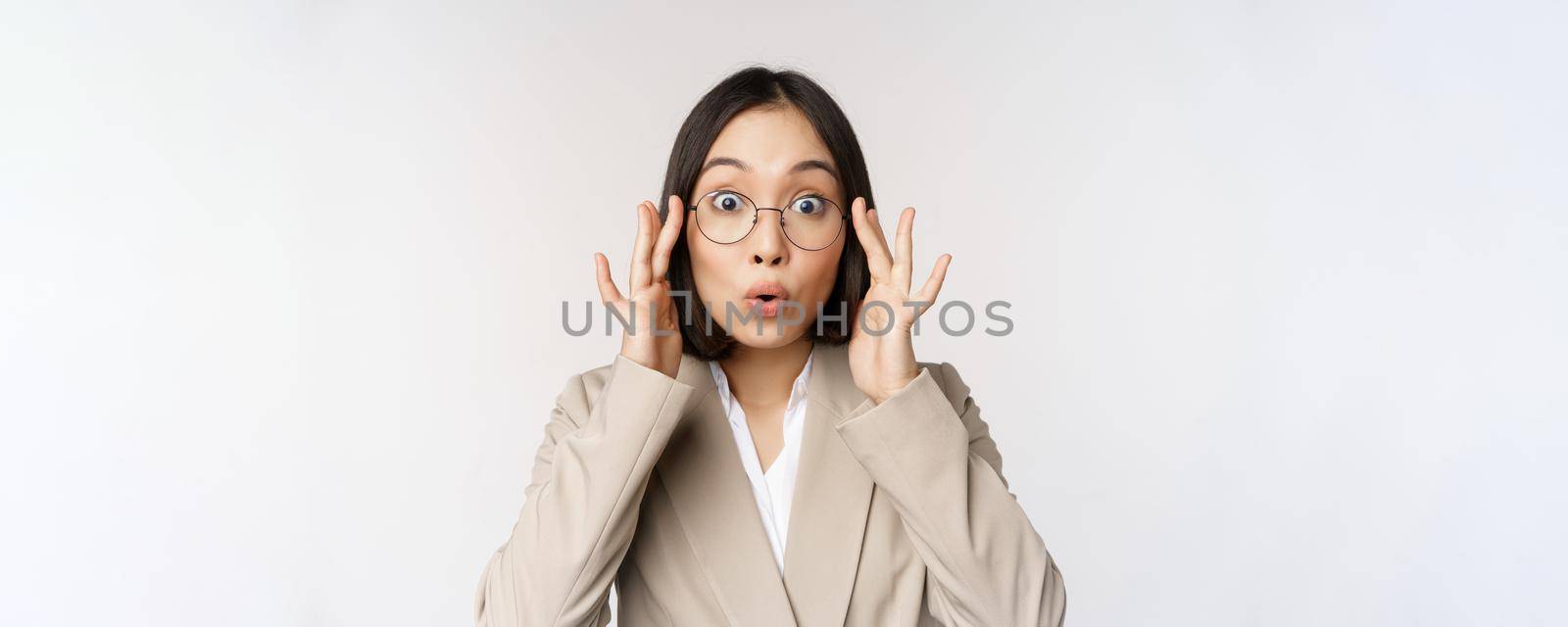 Portrait of asian businesswoman in glasses, looking surprised amazed at camera, standing in suit over white background by Benzoix