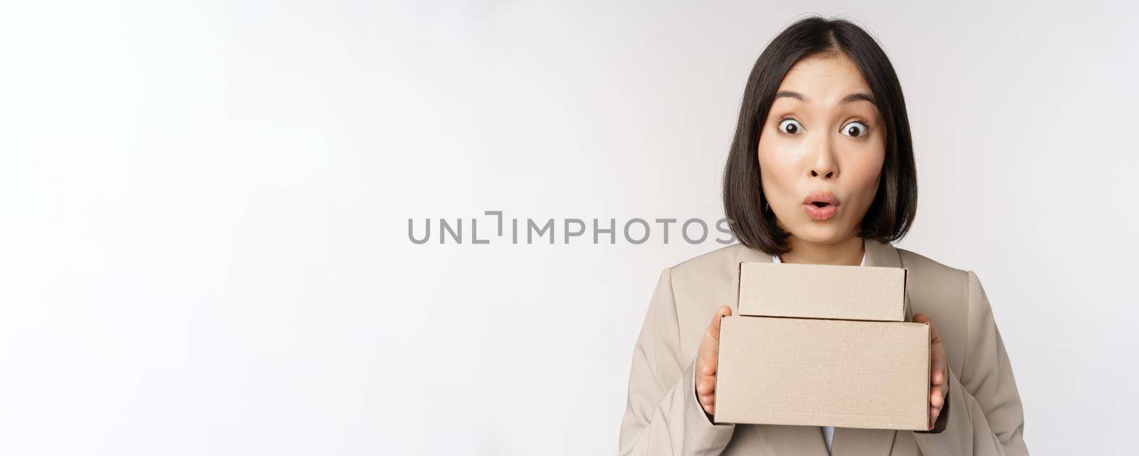 Portrait of asian saleswoman looking surprised, holding boxes, delivery goods, standing amazed in suit against white background.