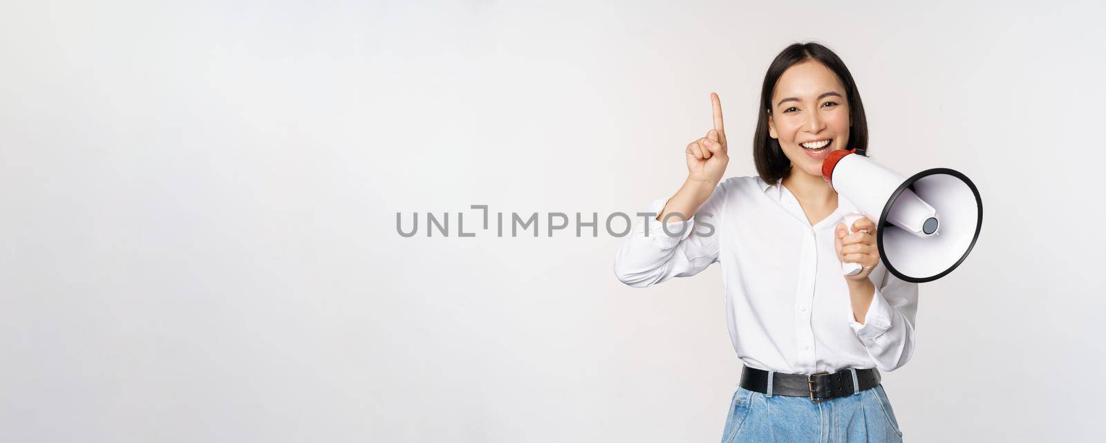 Smiling happy asian girl talking in megaphone and pointing up, announcing discount promo, showing advertisement on top, standing over white background.