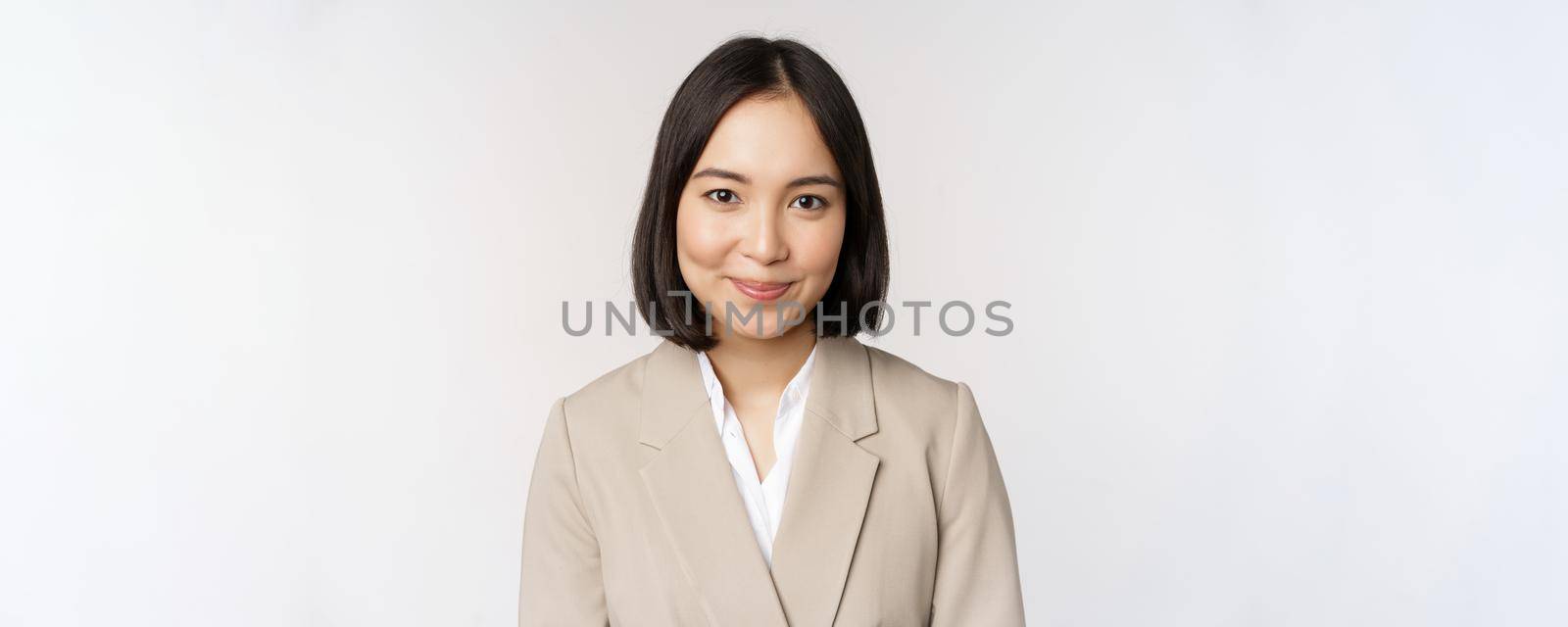 Close up portrait of businesswoman, asian female entrepreneur in suit, smiling and looking professional, standing against white background.