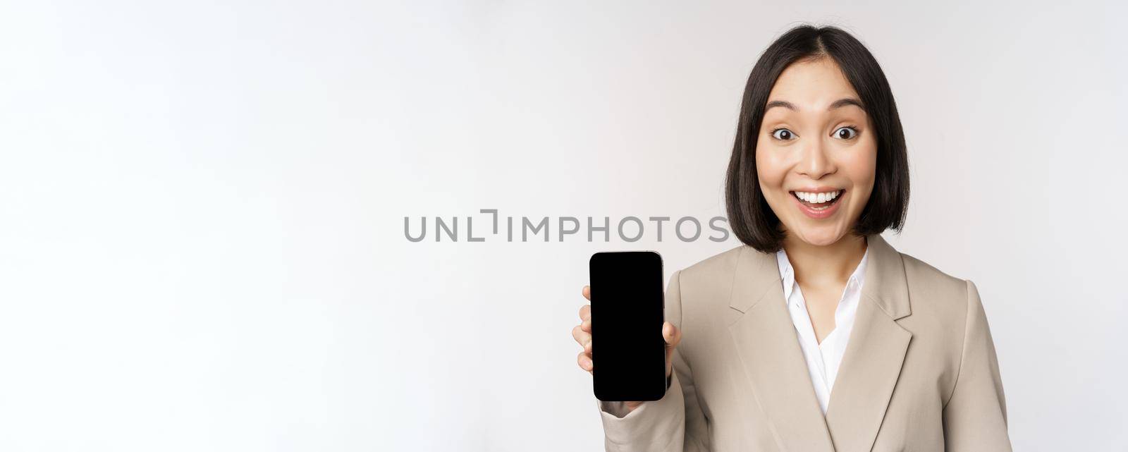 Image of asian corporate woman showing app interface, mobile phone screen, making surprised face expression, wow, standing over white background.