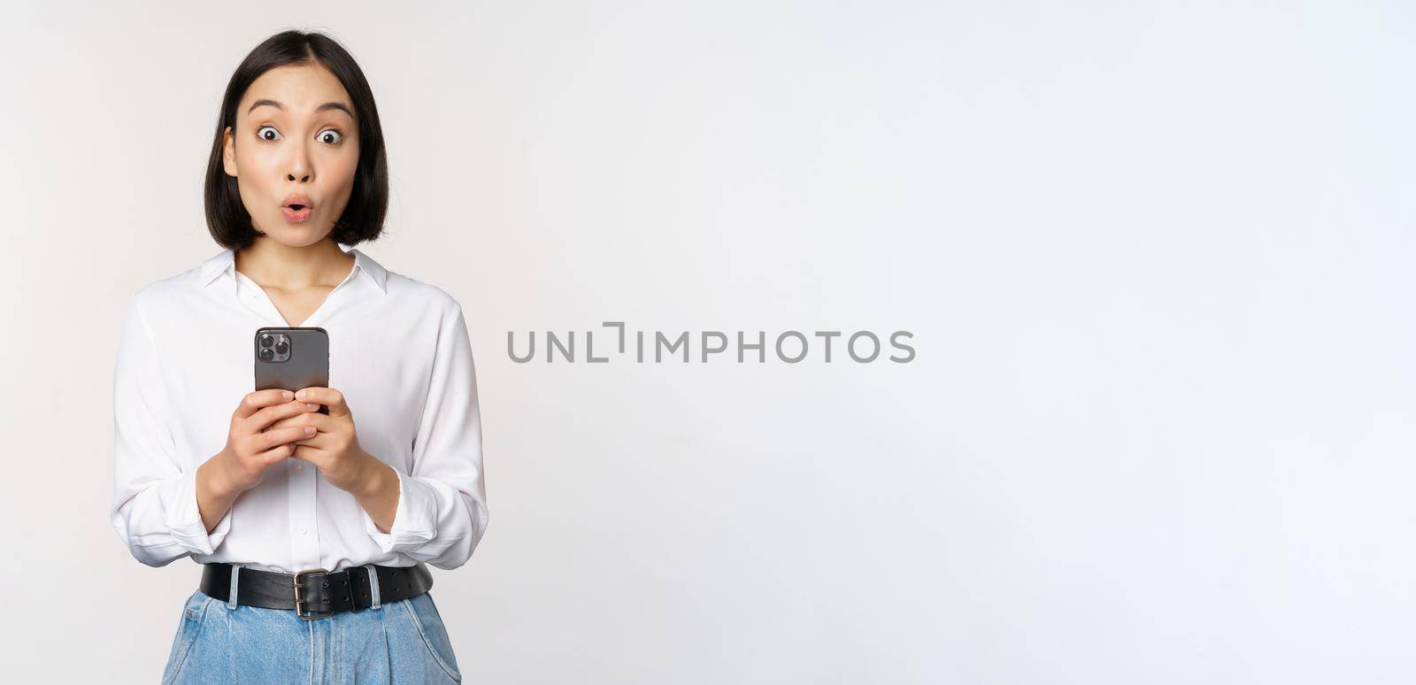 Technology concept. Portrait of asian woman with mobile phone, girl holding smartphone and reacting surprised at cellphone notification, app info, white background by Benzoix