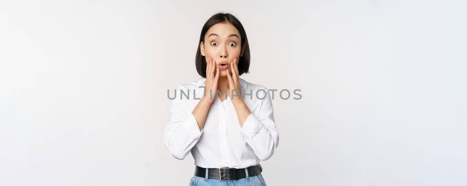 Image of asian female model looking surprised, staring amazed, reacting at surprise big news, standing over white background by Benzoix