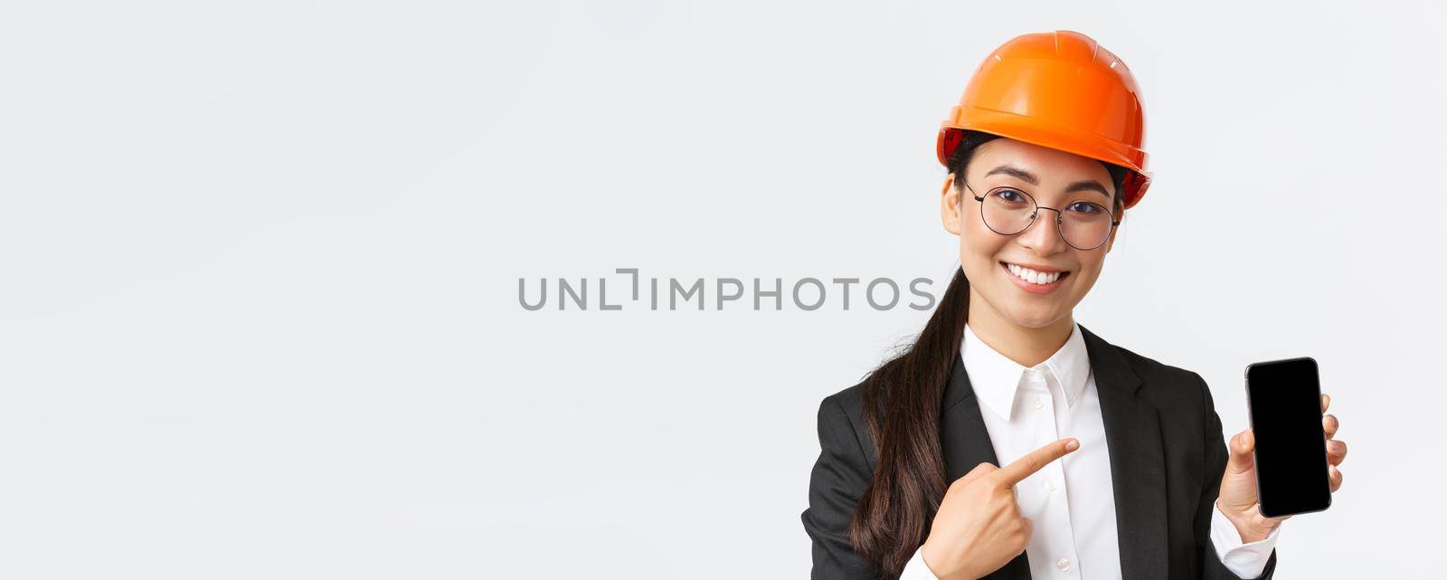 Close-up of smiling professional female asian construction engineer, architect in suit and safety mask introduce application, pointing finger at smartphone display, white background.