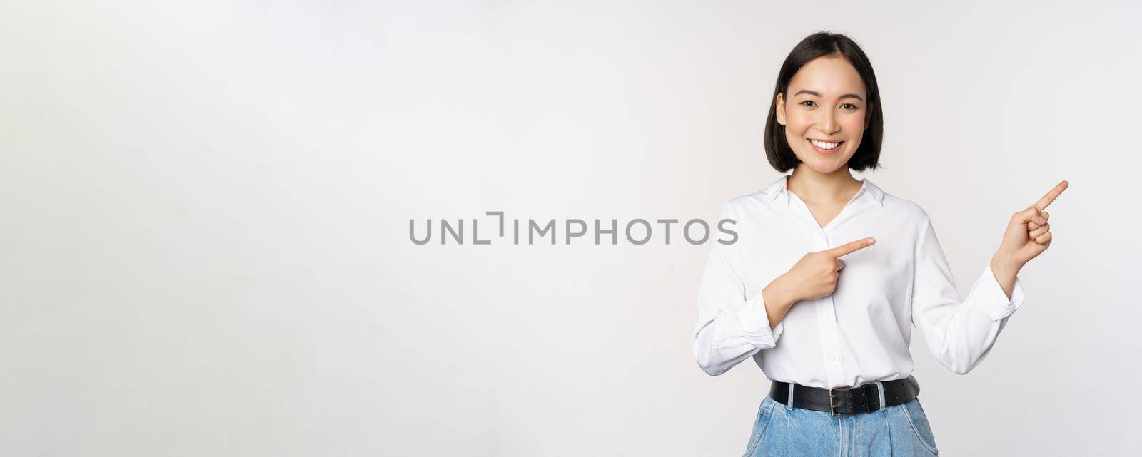 Portrait of beautiful asian woman, saleswoman pointing fingers right and showing info, banner or logo, sale advertisement, standing over white background by Benzoix