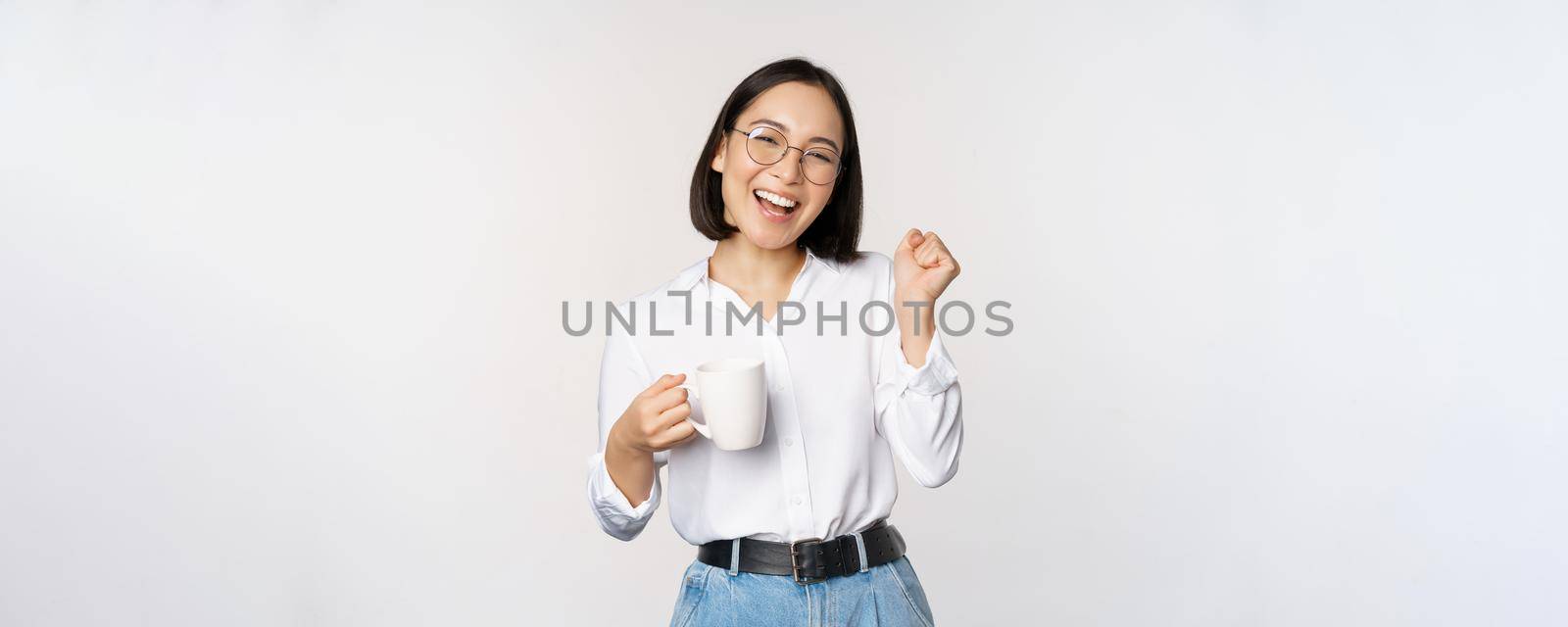 Happy dancing woman drinking coffee or tea from mug. Korean girl with cup, standing over white background by Benzoix