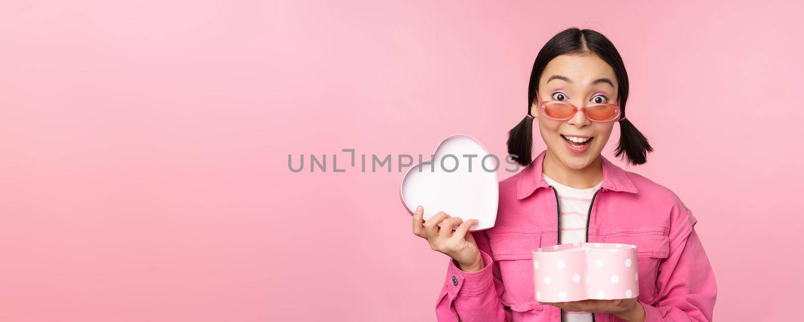 Cute asian girl giving you gift in heart shaped box, kissing and smiling, concept of holiday and celebration, standing over pink background by Benzoix