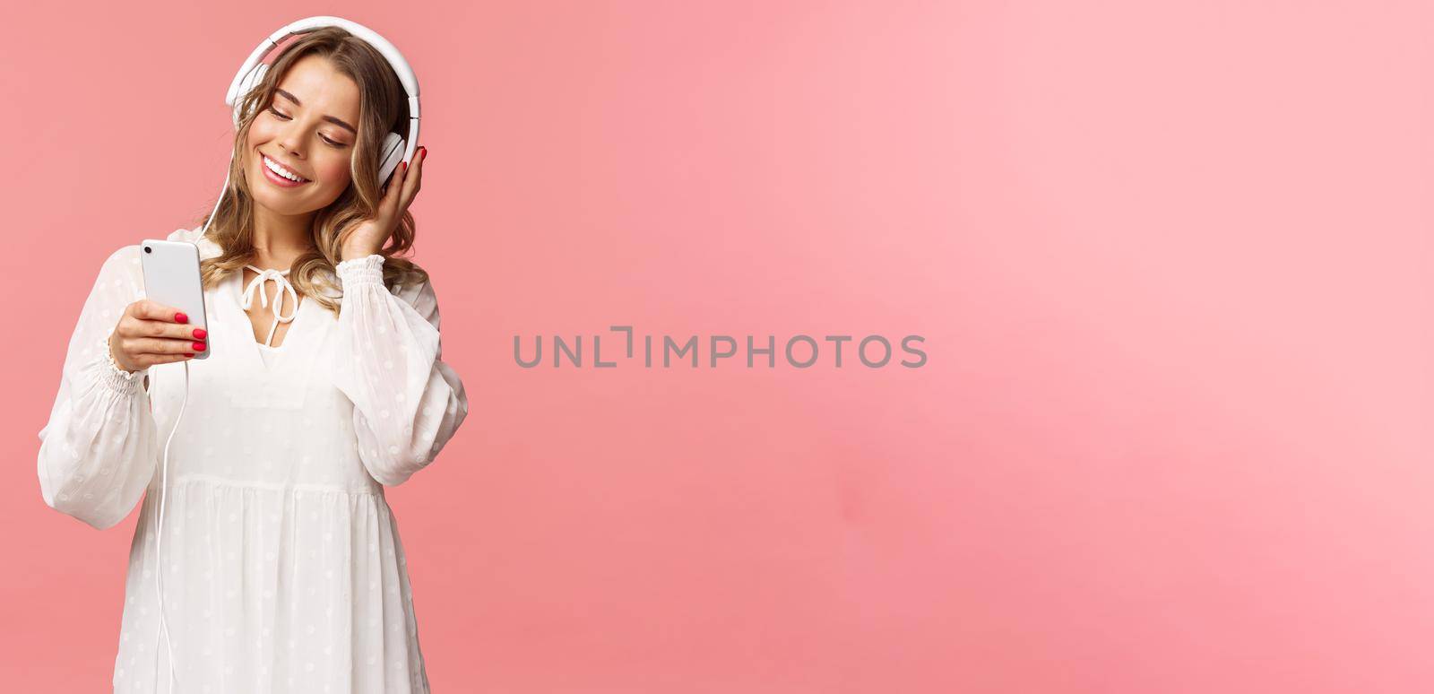 Portrait of dreamy cute and tender blond girl in white dress, holding smartphone, listen music in headphones, smiling at display mobile phone as picking playlist for spring mood, pink background by Benzoix