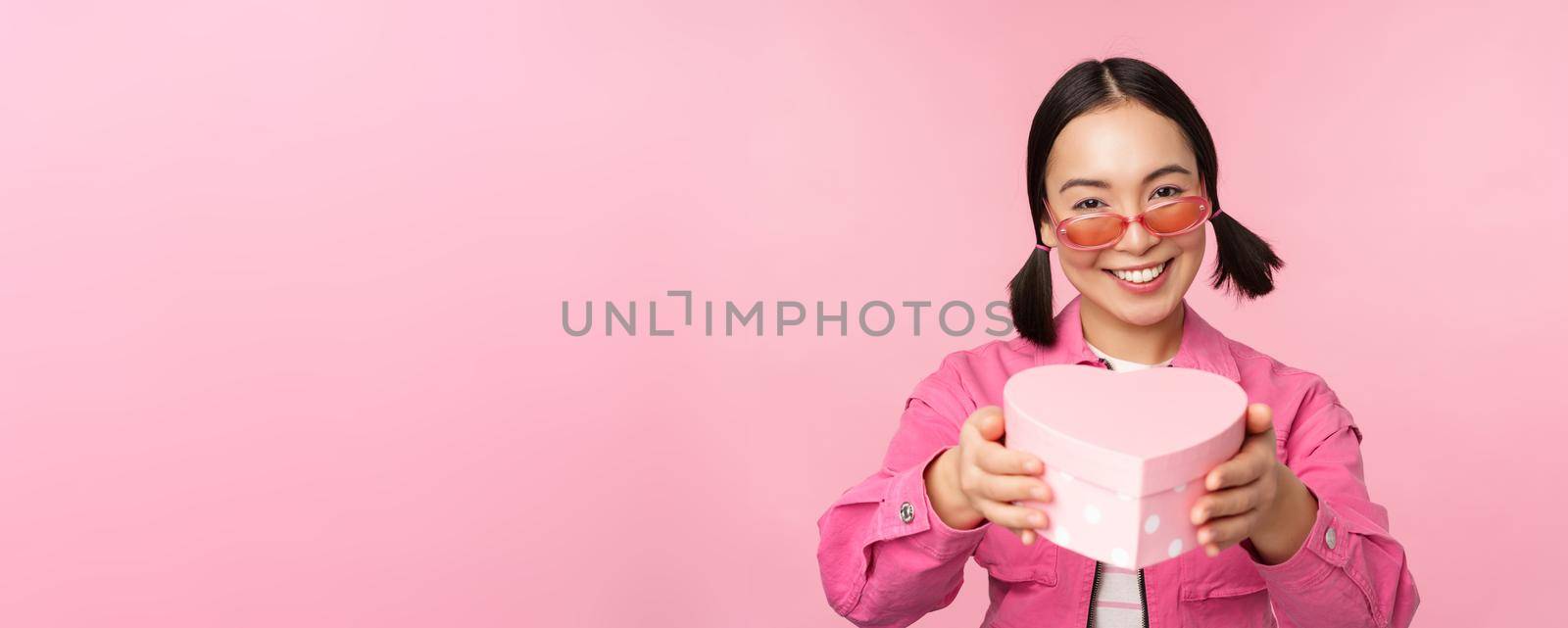 Cute asian girl giving you gift in heart shaped box, kissing and smiling, concept of holiday and celebration, standing over pink background by Benzoix