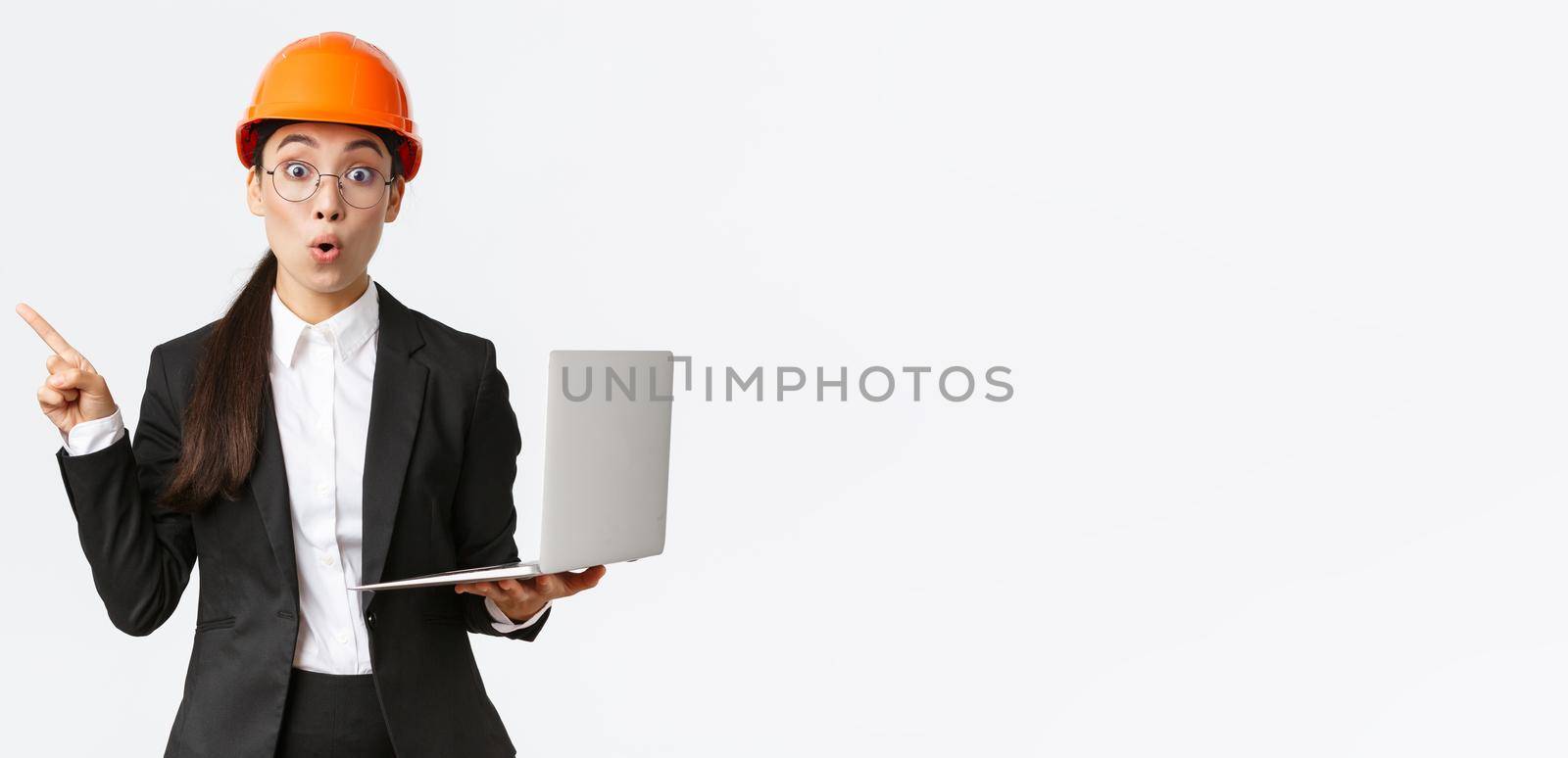 Impressed asian female entrepreneur at factory wearing safety helmet and business suit, pointing finger left and holding laptop computer, showing diagram at enterprise, white background by Benzoix