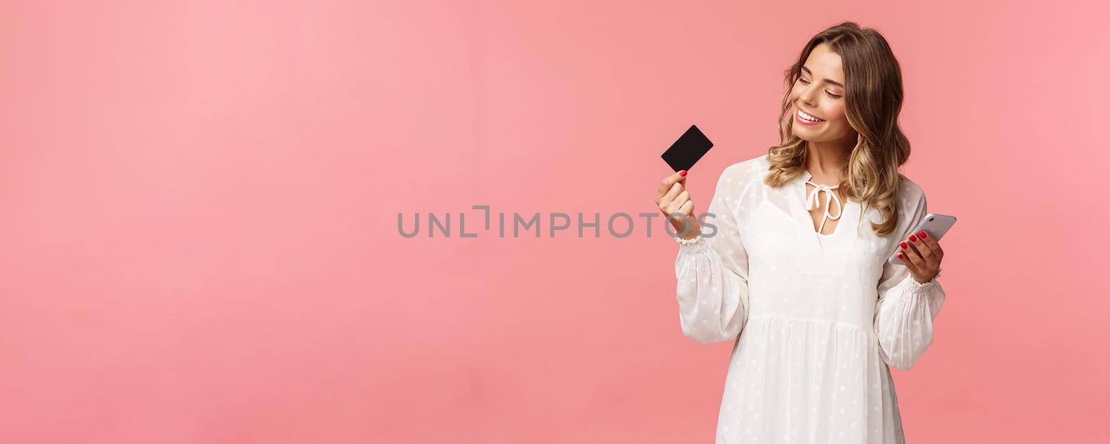 Portrait of happy good-looking young blond woman in white dress, looking pleased and satisfied at credit card, holding smartphone, recommend buy online, internet shopping concept.