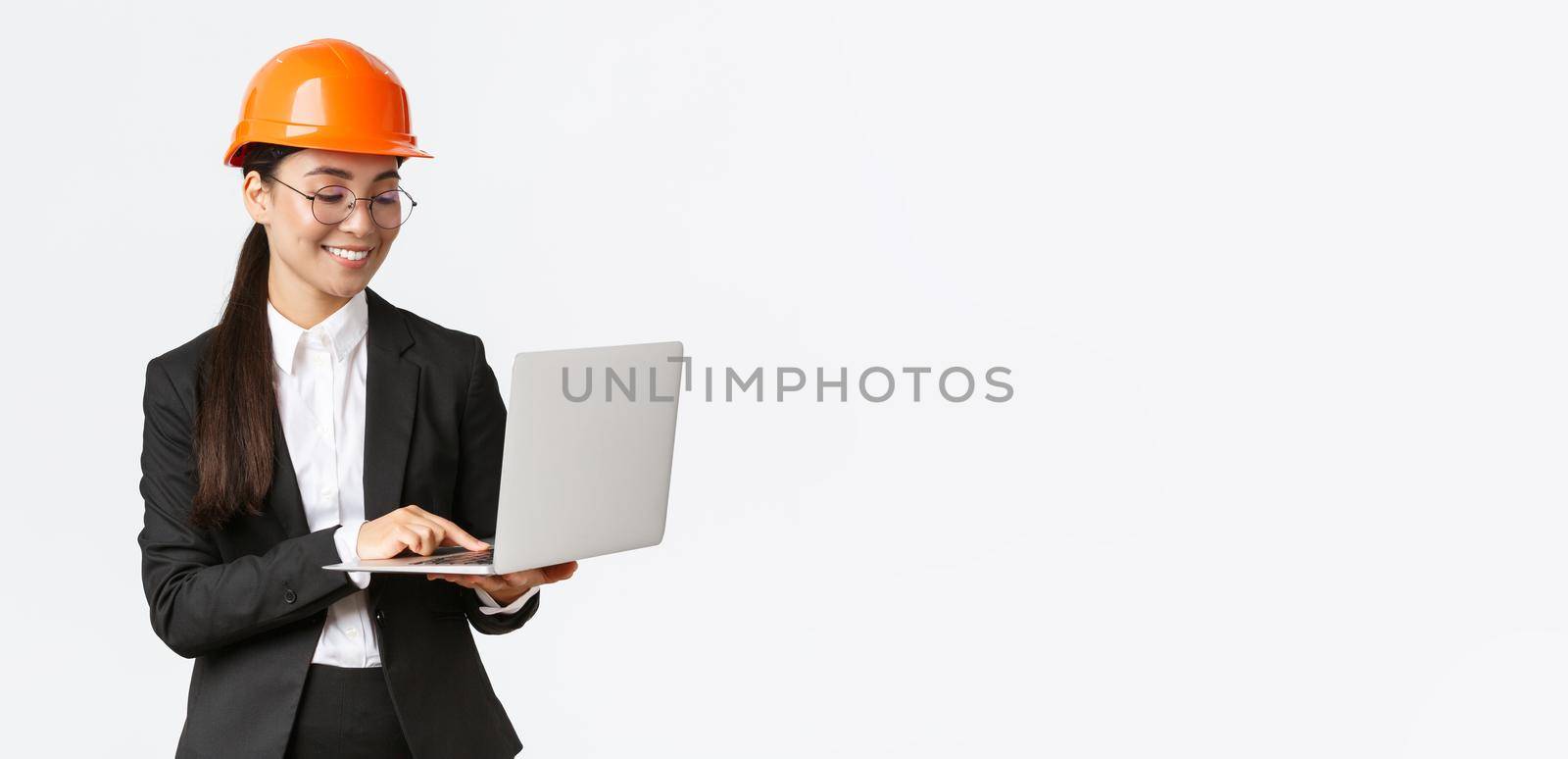 Smiling successful female asian industrial engineer, factory manager in safety helmet and business suit using laptop computer, checking with project or blueprints on screen.