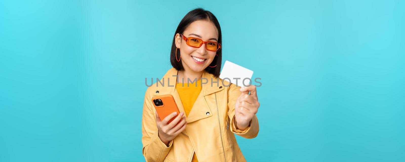 Online shopping. Stylish young asian woman in sunglasses, showing credit card and using smartphone, paying in internet, making purchase, standing over blue background.