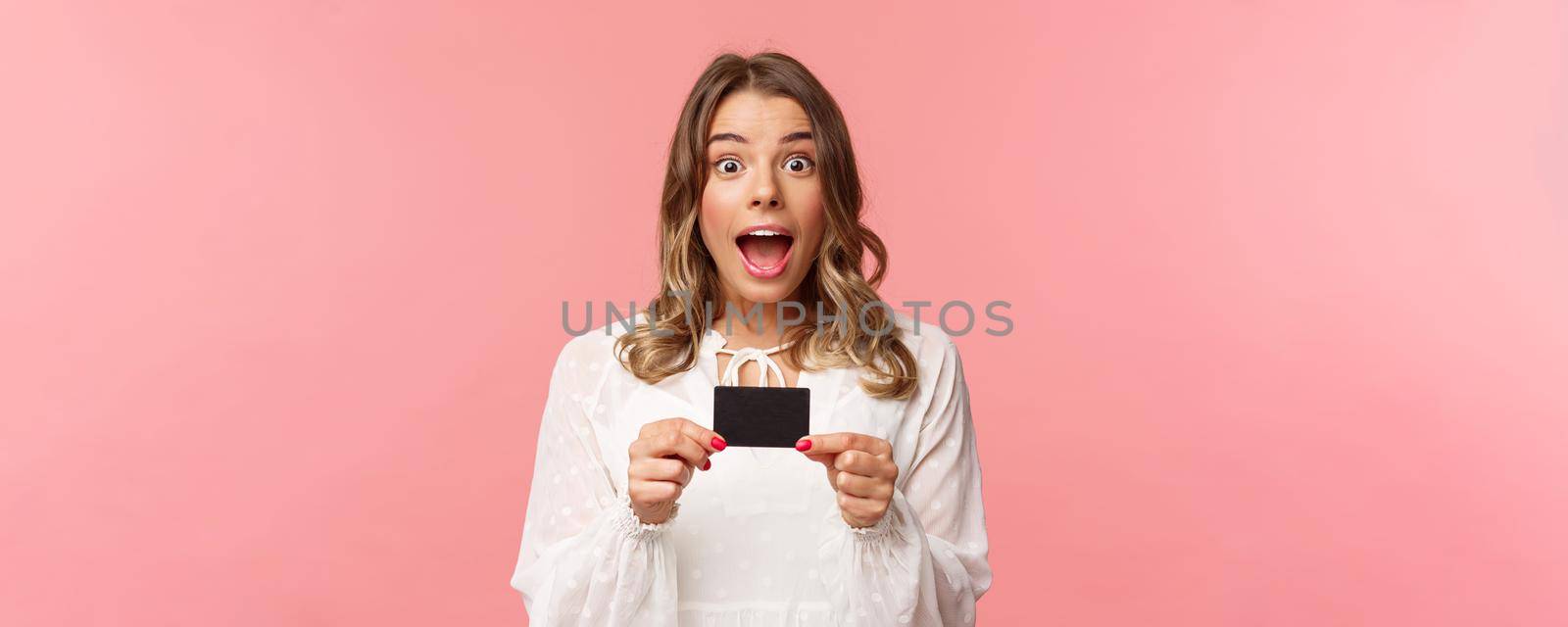Close-up portrait of emotive, excited and thrilled good-looking blond girl showing credit card and feeling very happy, telling about discounts, special offers, bank service, pink background.
