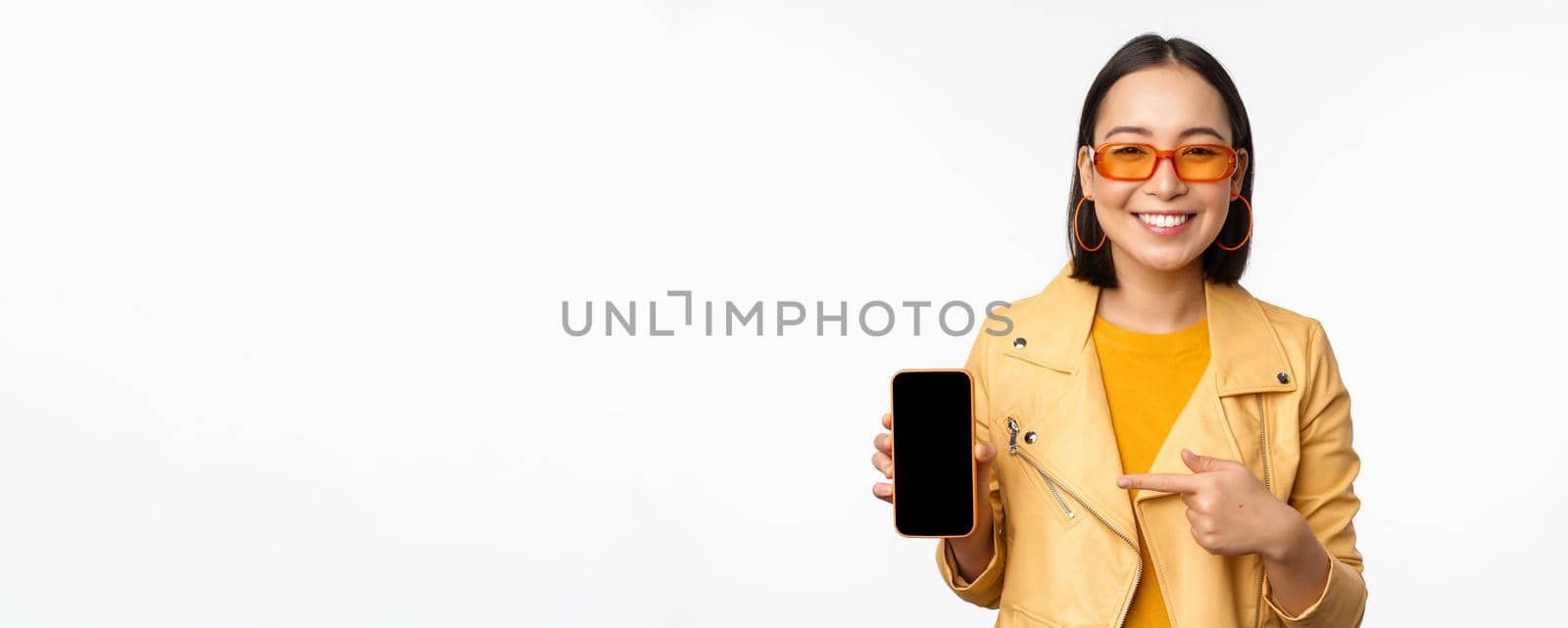 Portrait of stylish korean girl in sunglasses, smiling, pointing finger at smartphone screen, showing mobile phone application, standing over white background by Benzoix